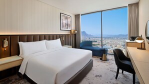 Una habitación de hotel moderna con una cama grande, un escritorio y vistas a un paisaje urbano a través de amplias ventanas.