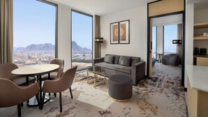 Una habitación de hotel moderna con un gran ventanal que ofrece vistas a la ciudad, un sofá, una mesa de comedor con sillas y un otomano pequeño y redondo.