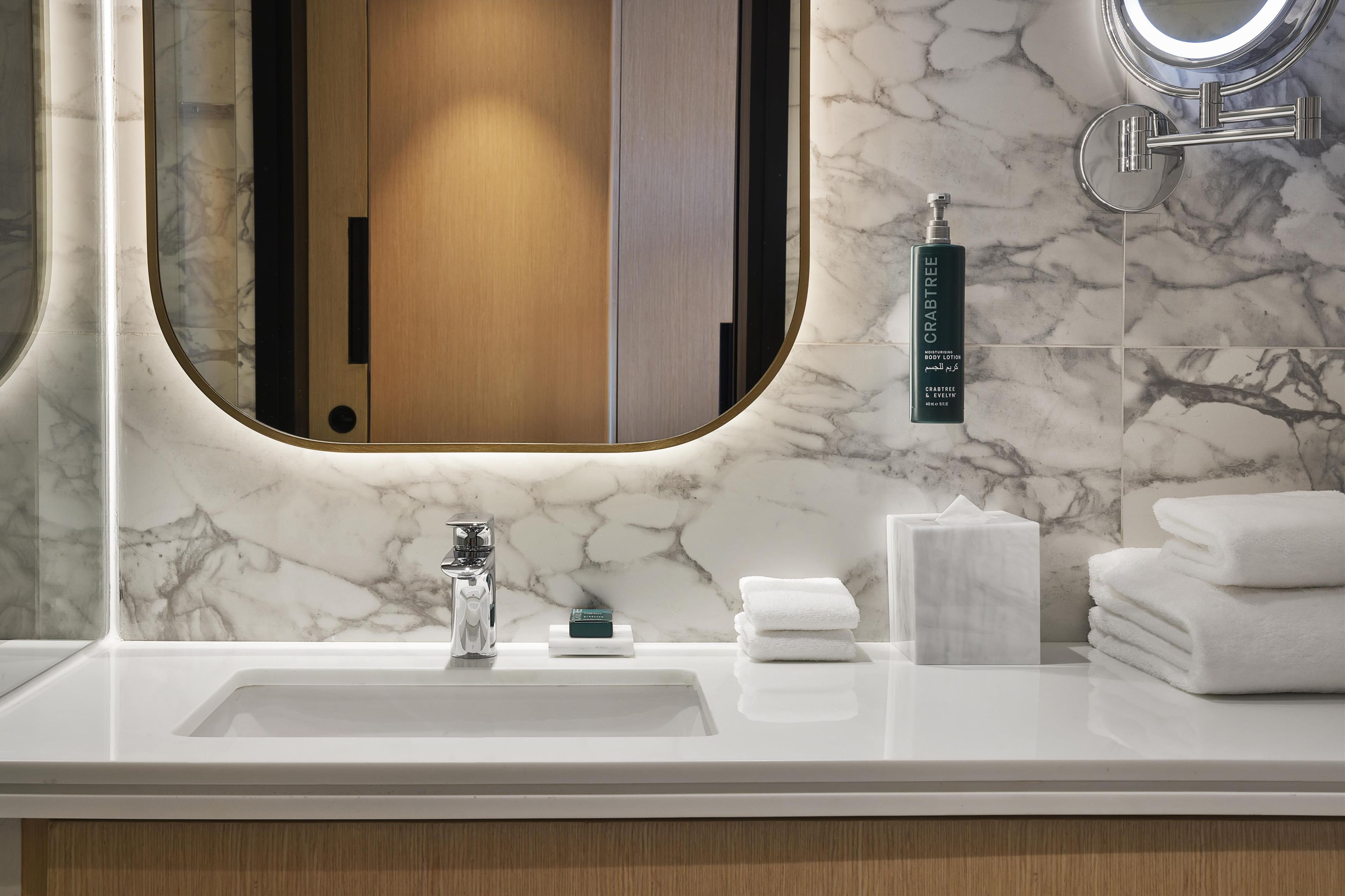 A bathroom with a marble sink, a mirror, a hand soap dispenser, and neatly stacked towels.