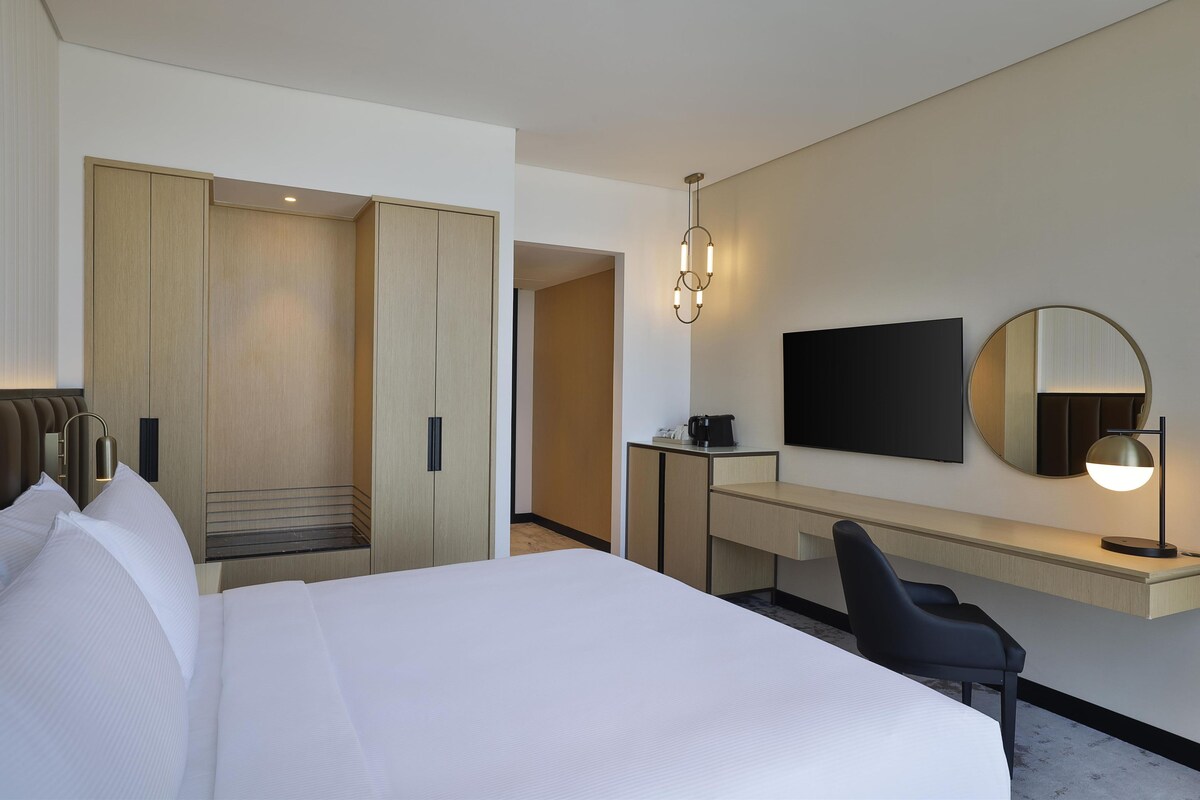 Habitación de hotel con una cama grande, un escritorio, televisor de pantalla plana, un espejo y una lámpara moderna.
