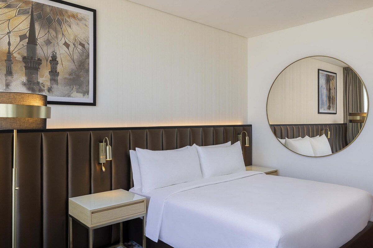 A hotel room with a large bed, a bedside table, a nightstand, a wall-mounted lamp, a round mirror, and a framed picture on the wall.
