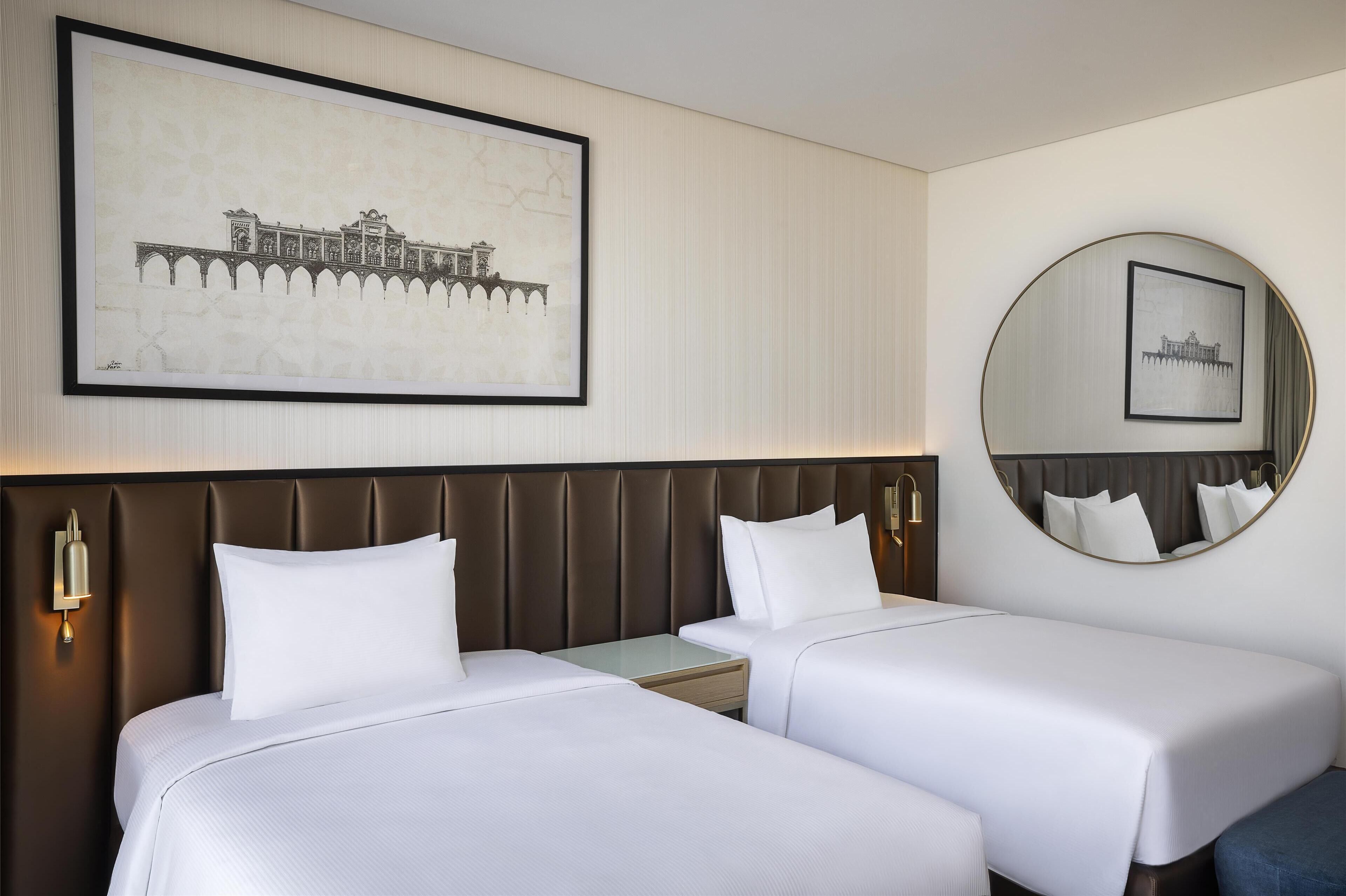 A hotel room with two beds, a large framed picture of a building, a round mirror, and a wall-mounted light fixture.