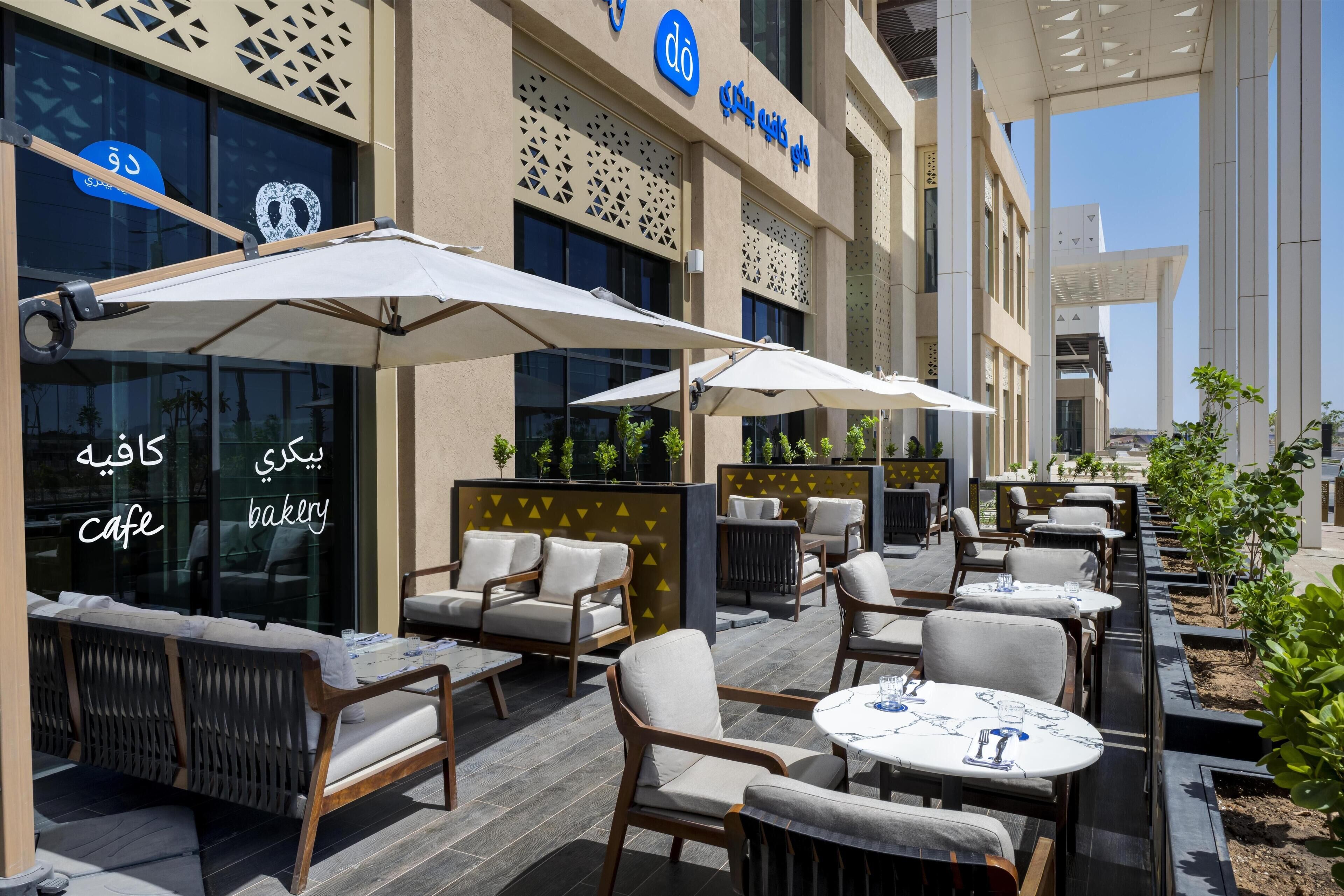 An outdoor cafe with seating and umbrellas, a bakery sign, and a modern building facade.