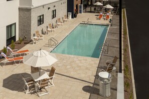 Un área de piscina en la azotea con sillones de descanso y sombrillas, un edificio de ladrillo y una piscina azul cristalina.