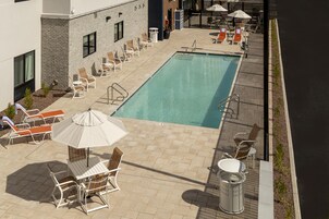 Un área de piscina en la azotea con sillones de descanso y sombrillas, un edificio de ladrillo y una piscina azul cristalina.