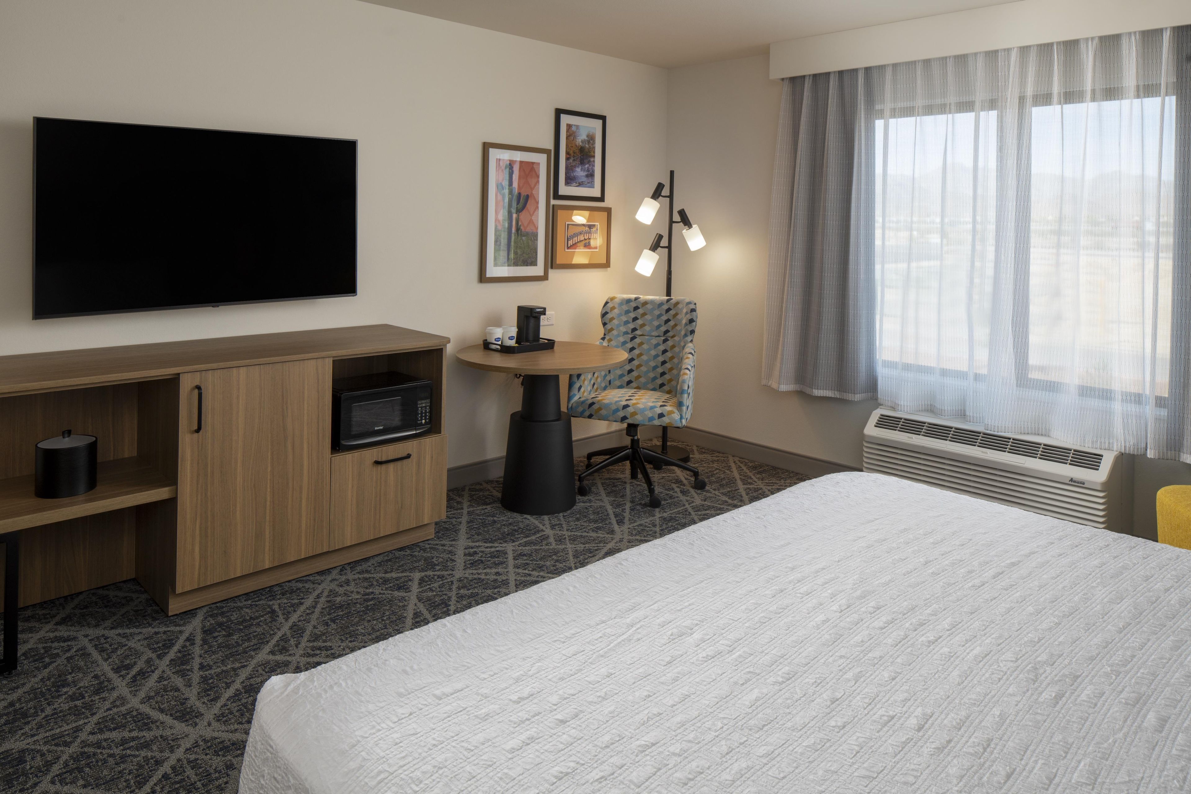 A hotel room with a bed, a TV, a desk with a microwave, a chair, and a window with curtains.