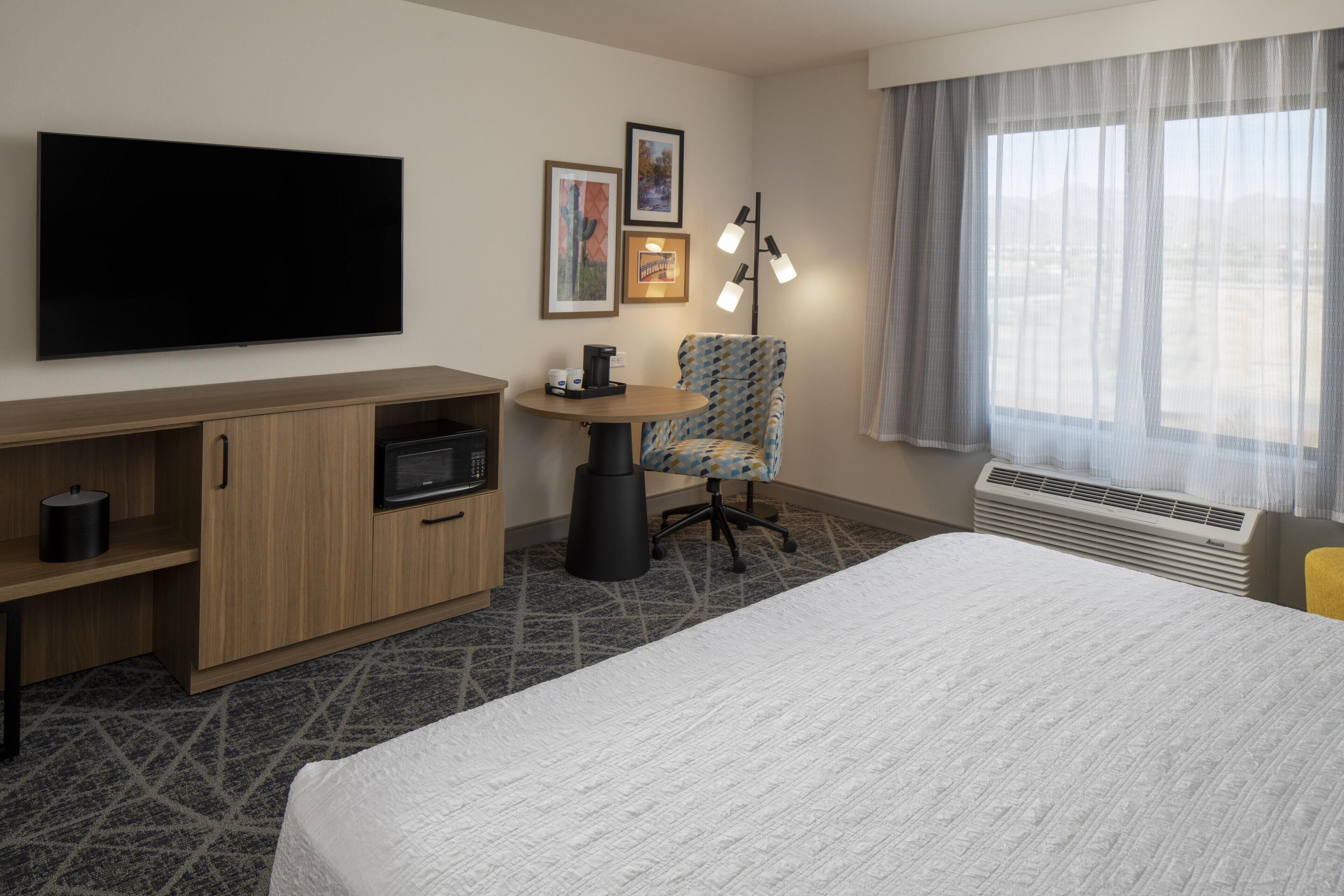 A hotel room with a bed, a TV, a desk with a microwave, a chair, and a window with curtains.