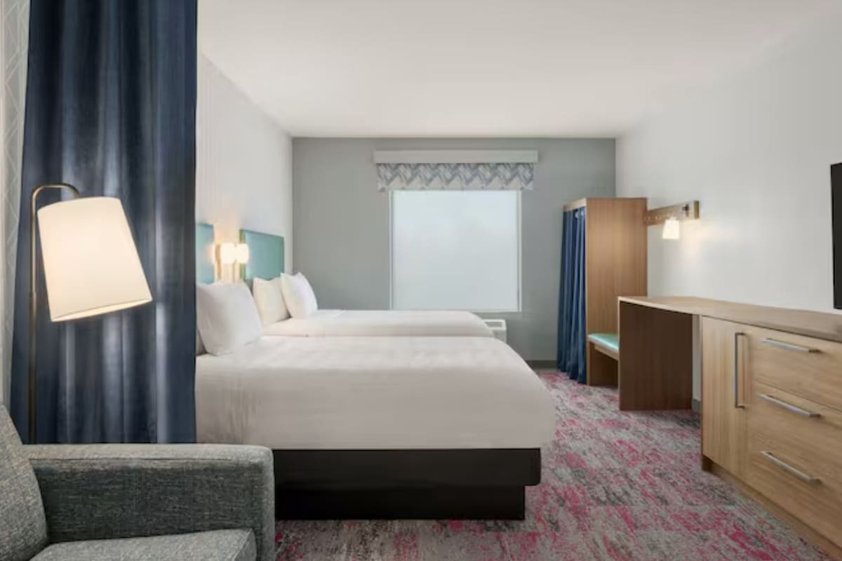 A hotel room with a bed, a desk, a chair, a lamp, and a television.