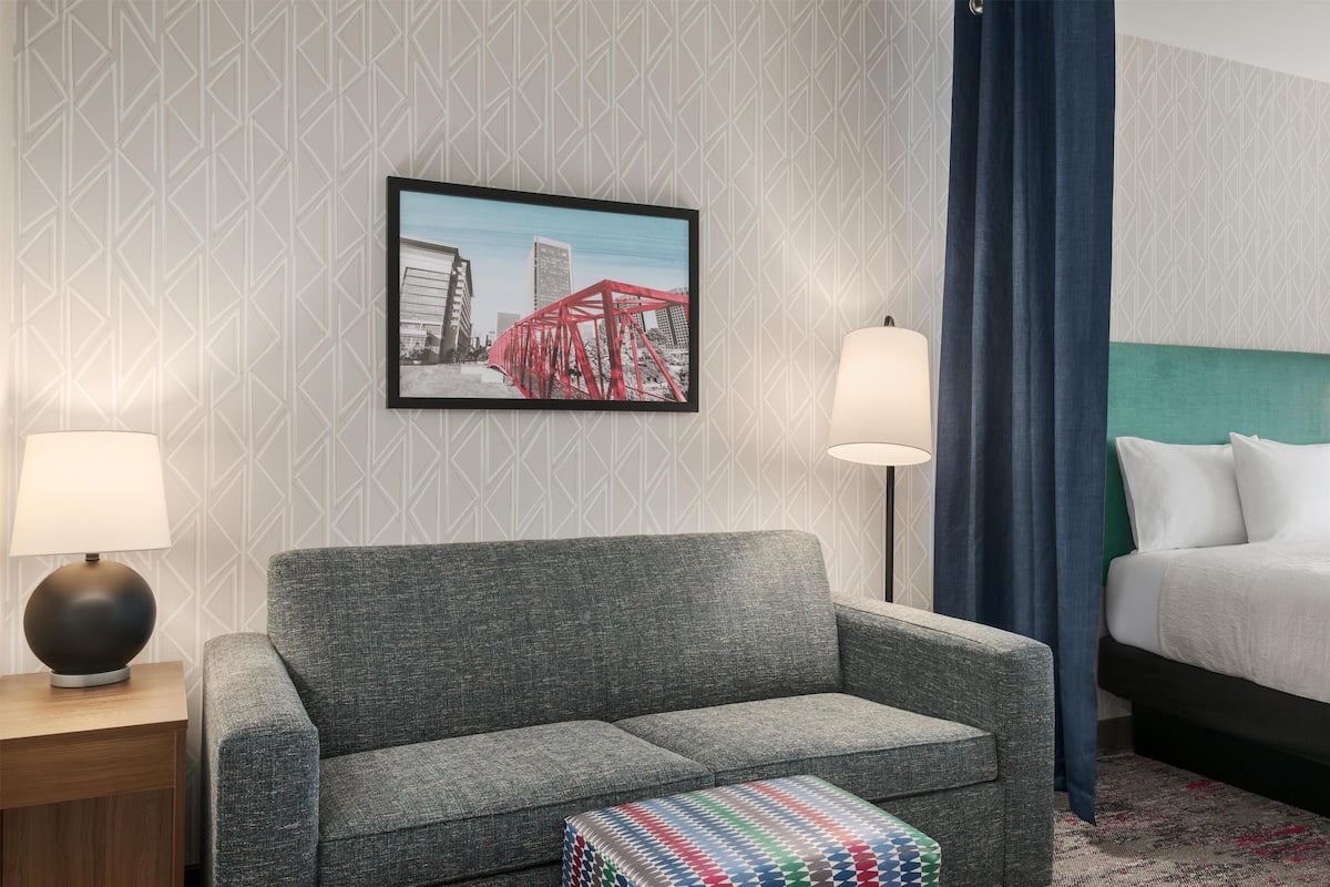 A hotel room with a grey sofa, a small ottoman, a framed picture of a bridge, a bed with white linens, and a blue curtain.