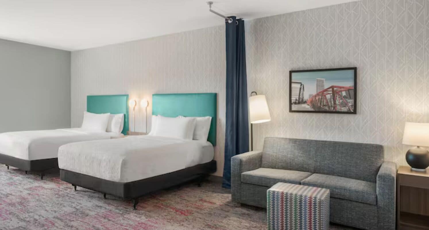 Habitación de hotel con dos camas, un sofá, una alfombra estampada y un cuadro de un paisaje urbano en la pared.