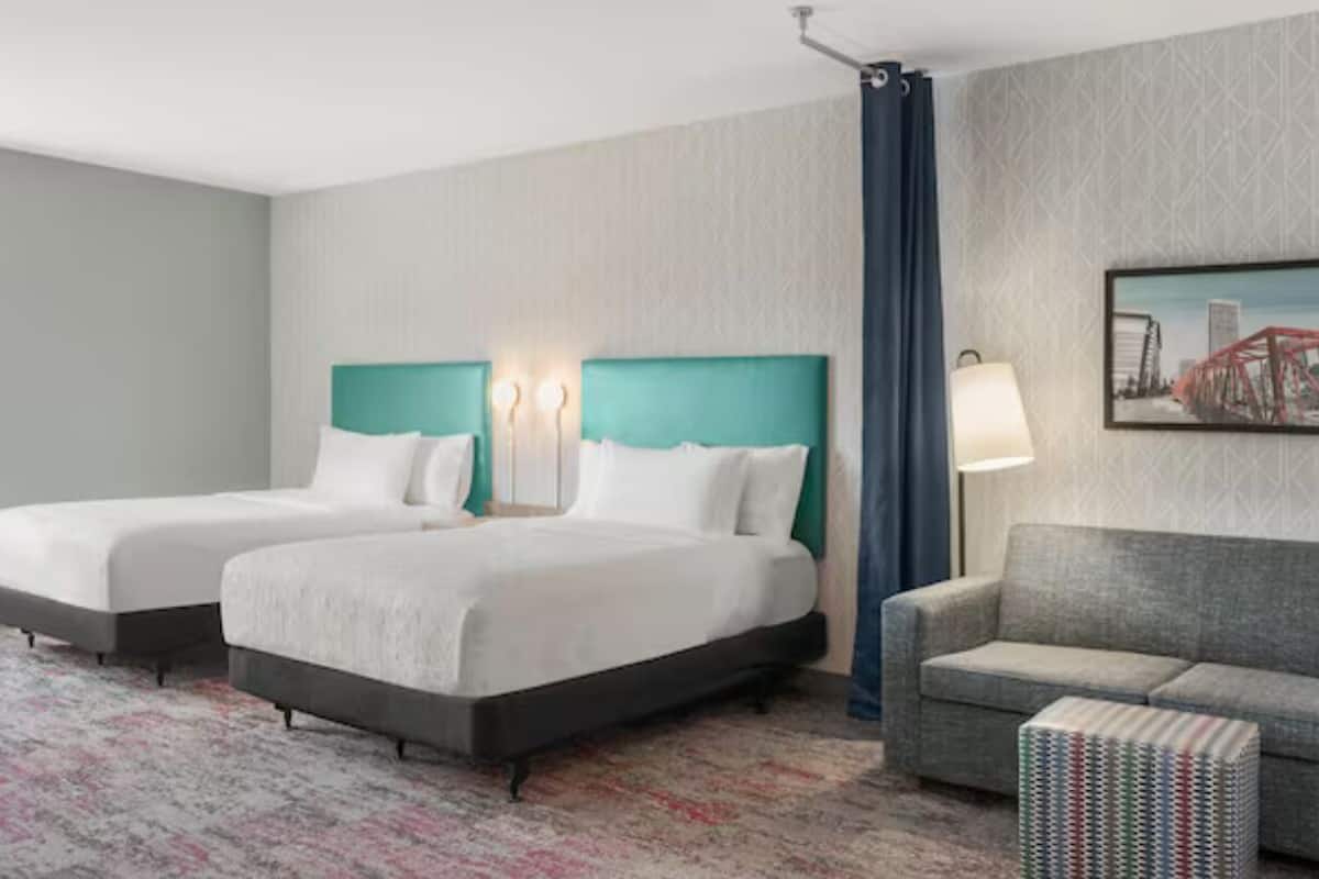 A hotel room with two beds, a sofa, a patterned carpet, and a cityscape painting on the wall.
