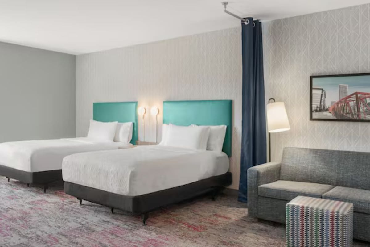 A hotel room with two beds, a sofa, a patterned carpet, and a cityscape painting on the wall.