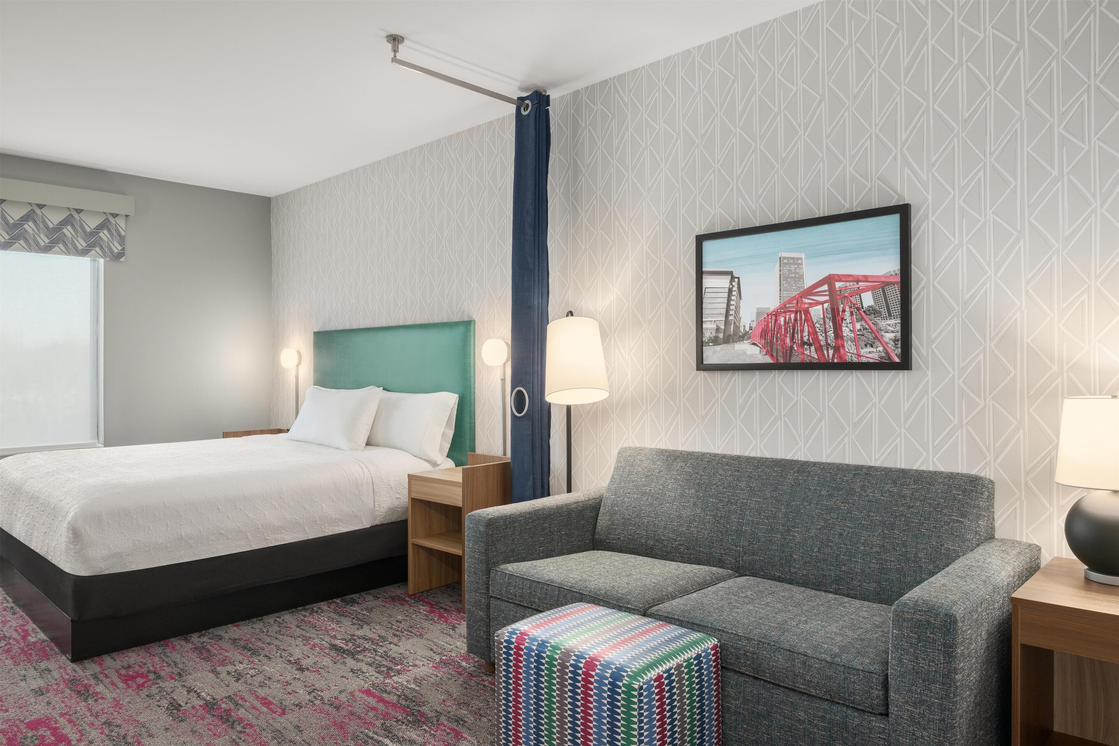 Un cuarto de hotel con una cama, un sofá gris, un reposapiés colorido, una mesita de noche, una lámpara y un cuadro en la pared.