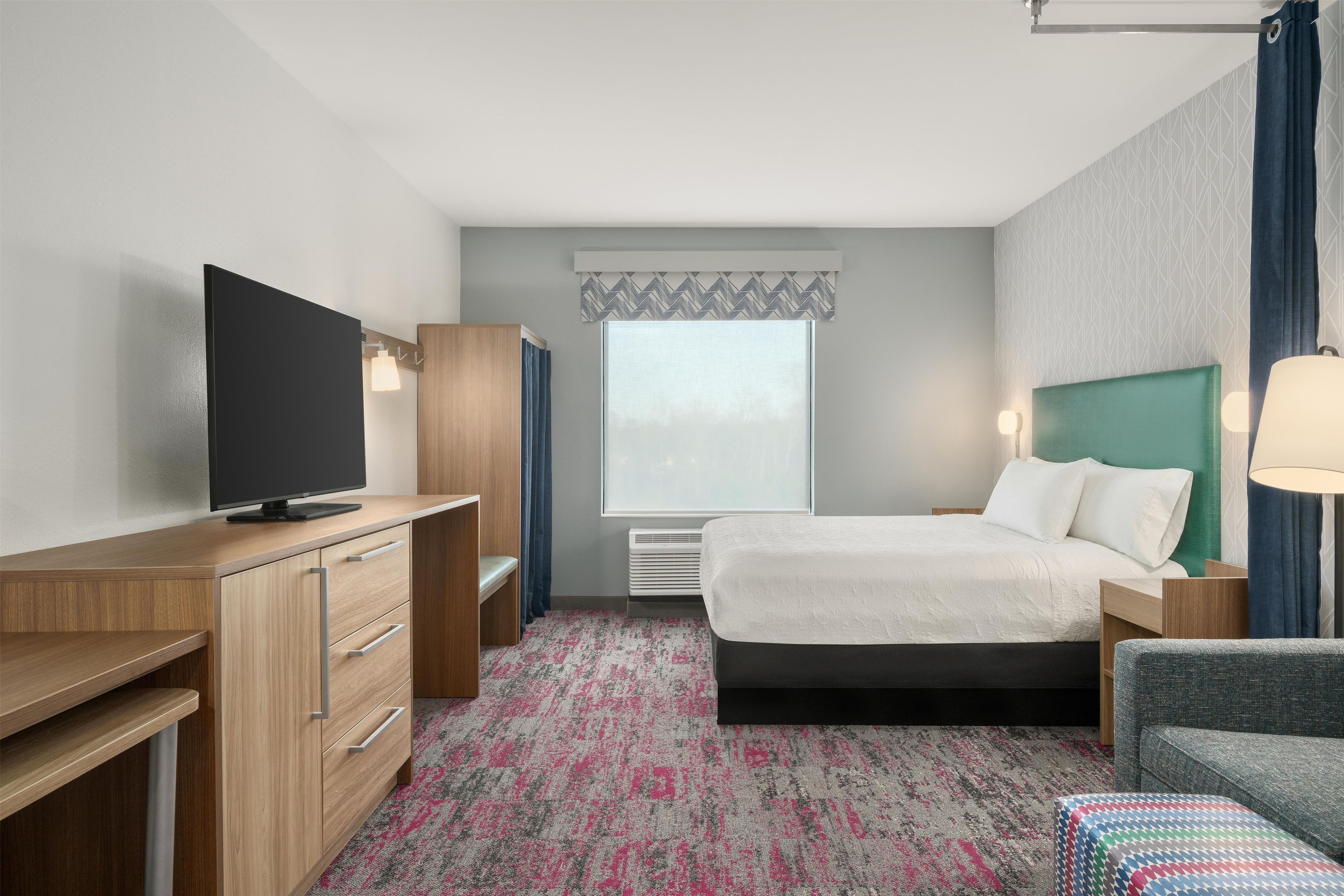 Ein Hotelzimmer mit Bett, Schreibtisch mit Fernseher, Kleiderschrank, Fenster mit Vorhängen und einem gemusterten Teppich.
