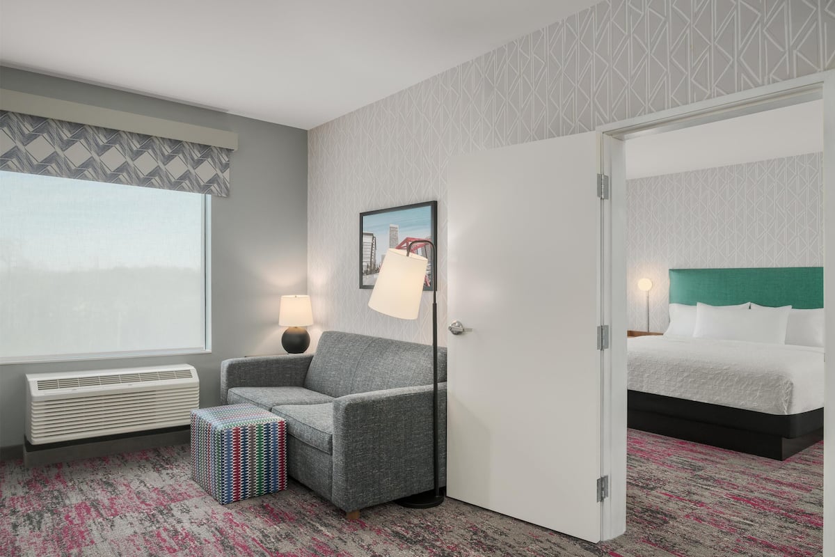 A hotel room with a grey sofa, a small ottoman, a bed with white bedding, a framed picture on the wall, and a window with a view.