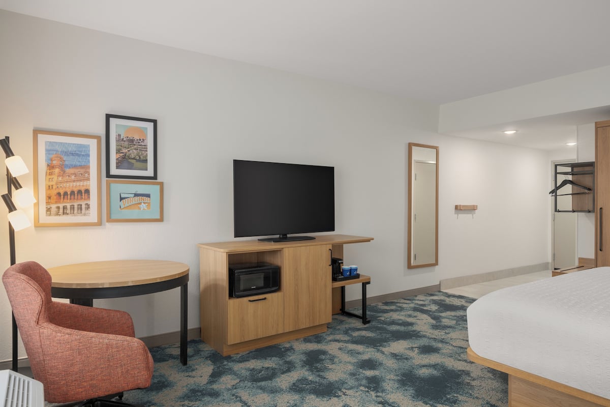 A hotel room with a bed, a TV on a wooden cabinet, a chair, a small table, and framed pictures on the wall.
