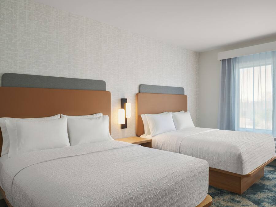A hotel room with two beds, a nightstand, a wall-mounted lamp, and a large window with curtains.