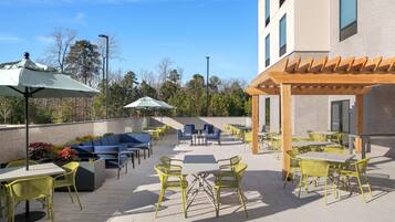 An outdoor patio with tables and chairs, a pergola, and a building in the background.