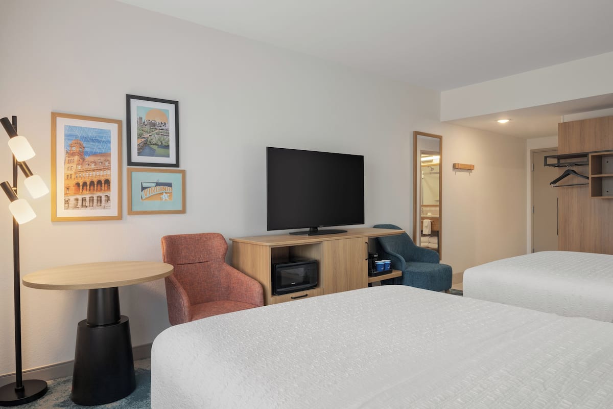 A hotel room with two beds, a TV, a desk, a chair, and framed pictures on the wall.