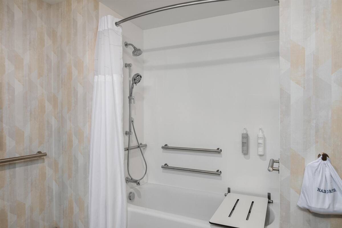 A shower area with a curved showerhead, a handheld showerhead, and a handheld shower attachment. There are two towel bars and a white towel rack. A white bag with 'HAIRDRY' is hanging on the wall.