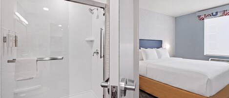 A hotel room with a glass-enclosed shower, a bed with a headboard, and a wall-mounted air conditioning unit.