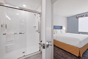 A hotel room with a glass-enclosed shower, a bed with a headboard, and a wall-mounted air conditioning unit. - Home2 Suites By Hilton Raleigh Crabtree (Raleigh)