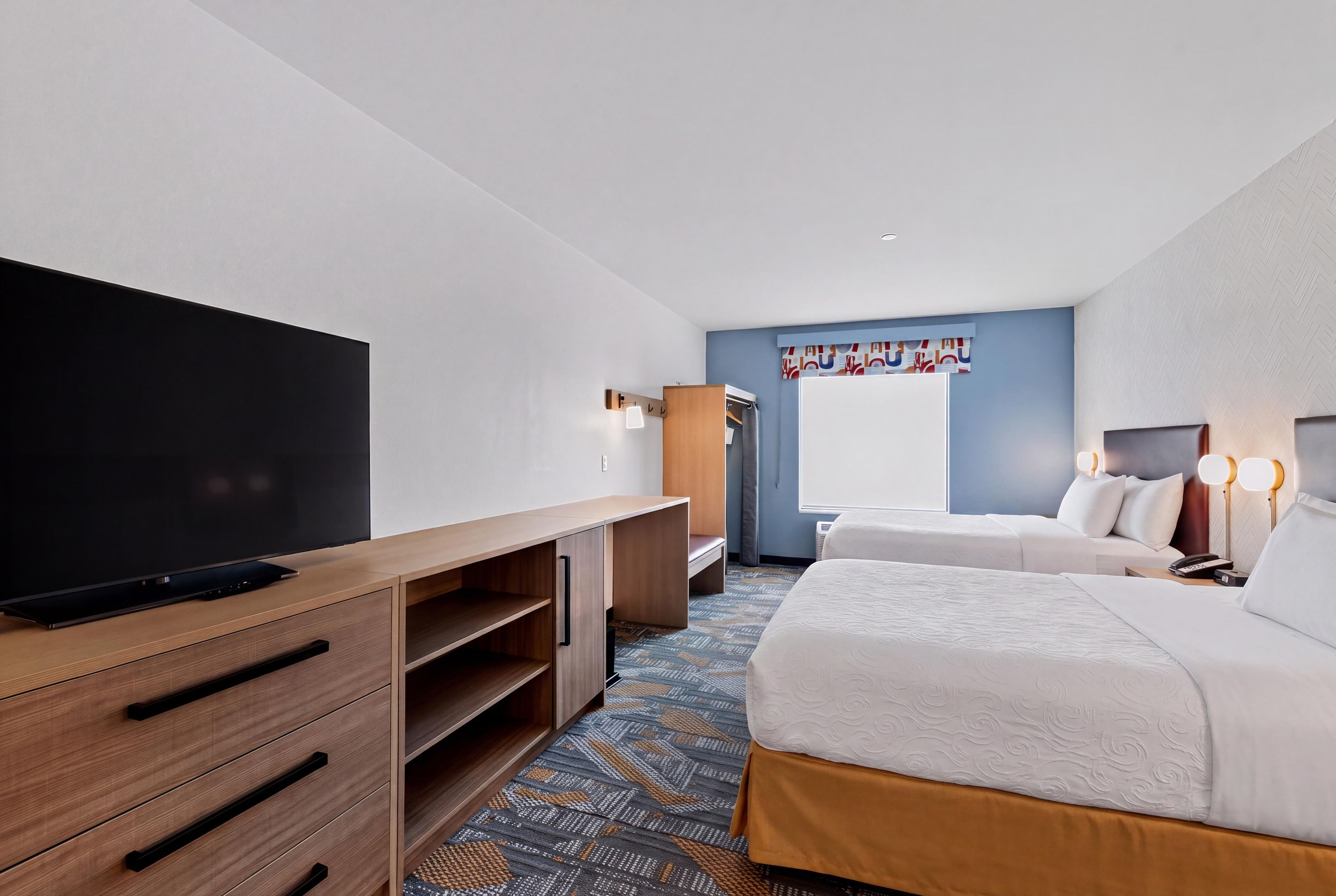 A hotel room with a flat-screen TV, wooden furniture, a bed with white bedding, and a patterned carpet.