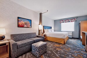 A hotel room with a sofa, a bed, a nightstand, a lamp, and a patterned carpet. - Home2 Suites By Hilton Raleigh Crabtree (Raleigh)