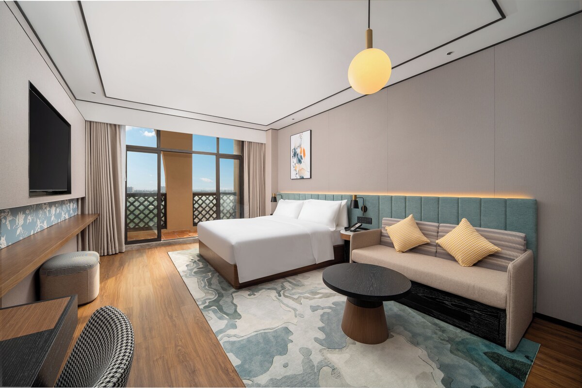 A modern hotel room with a bed, sofa, TV, and a view of the outdoors.