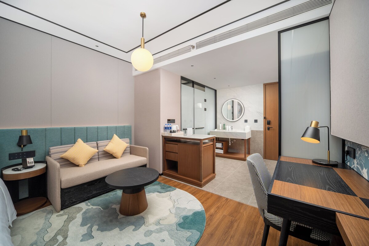 A modern hotel room with a sofa, round coffee table, dining table, and a small kitchen area.