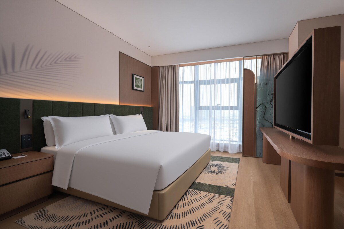 Una habitación de hotel moderna con una cama grande, un televisor de pantalla plana, una mesita de noche y un ventanal con cortinas.