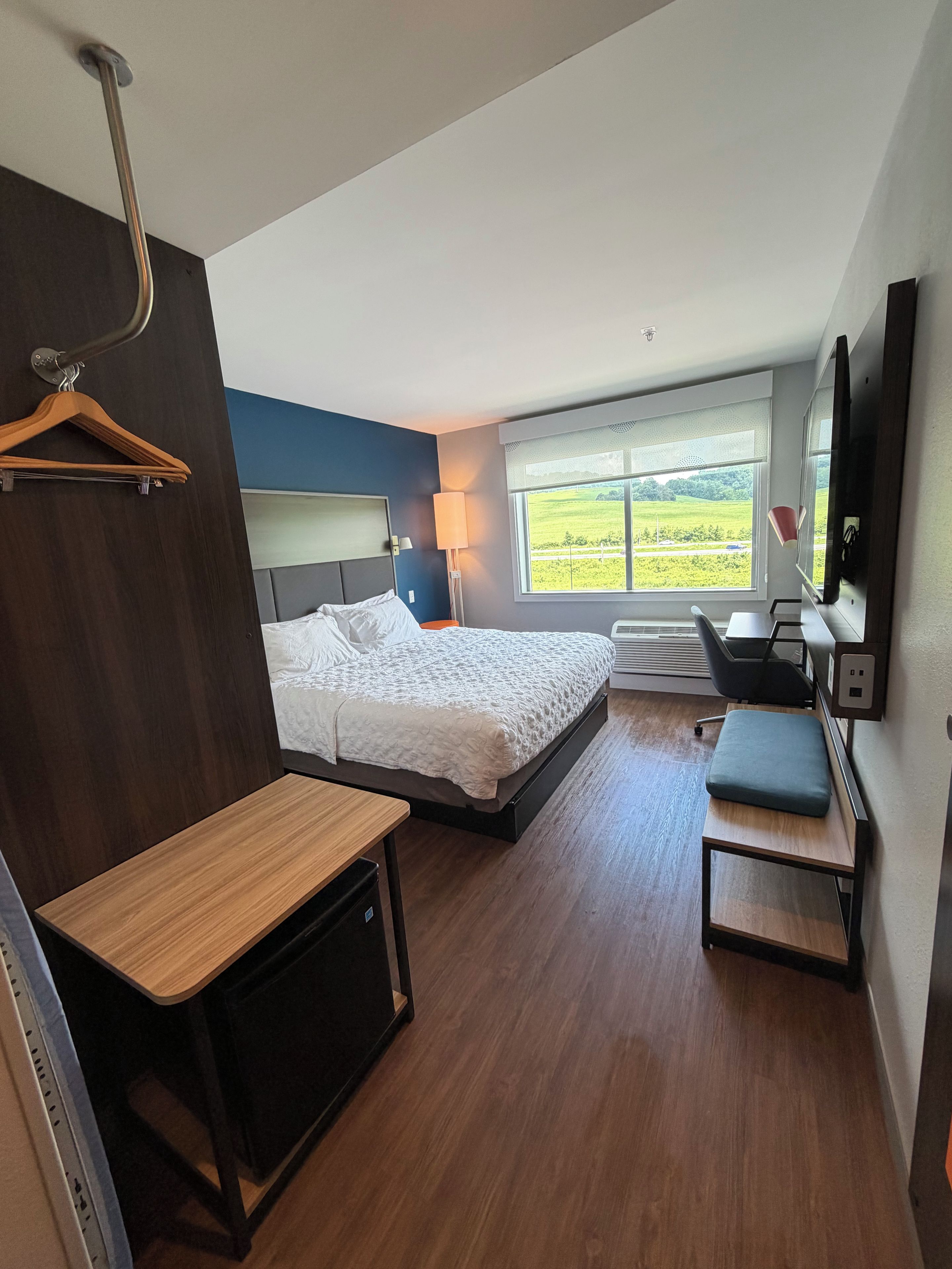 A modern hotel room with a bed, a desk, a chair, a TV, and a window with a view of the countryside.