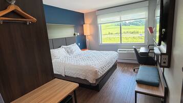 A modern hotel room with a bed, a desk, a chair, a TV, and a window with a view of the countryside.