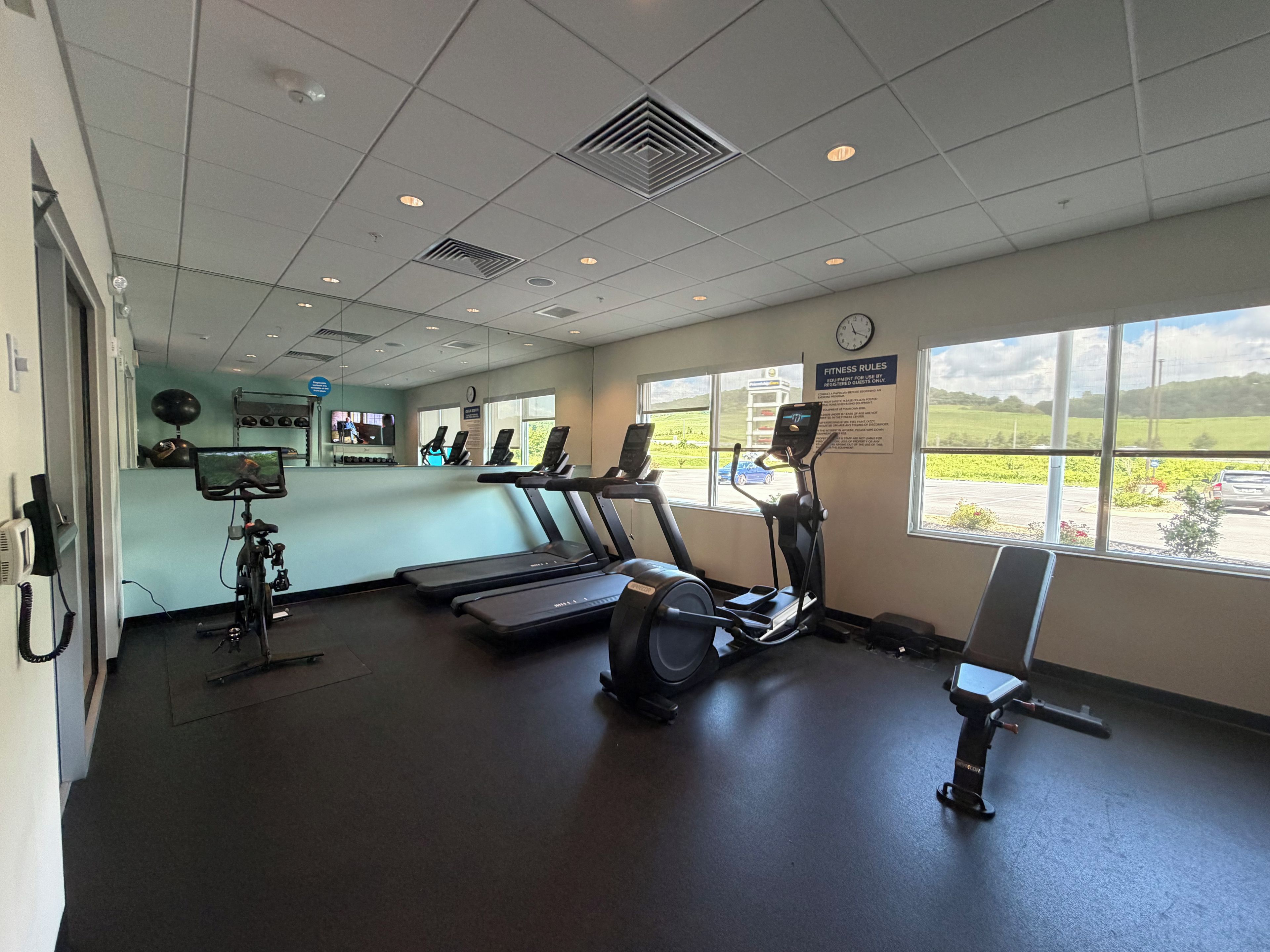 A gym with treadmills, an elliptical, and exercise bikes. There are windows with a view of a parking lot and greenery.
