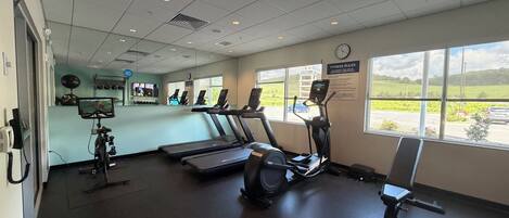 Un gimnasio con cintas de correr, elípticas y bicicletas estáticas. Hay ventanas con vistas a un estacionamiento y zonas verdes.