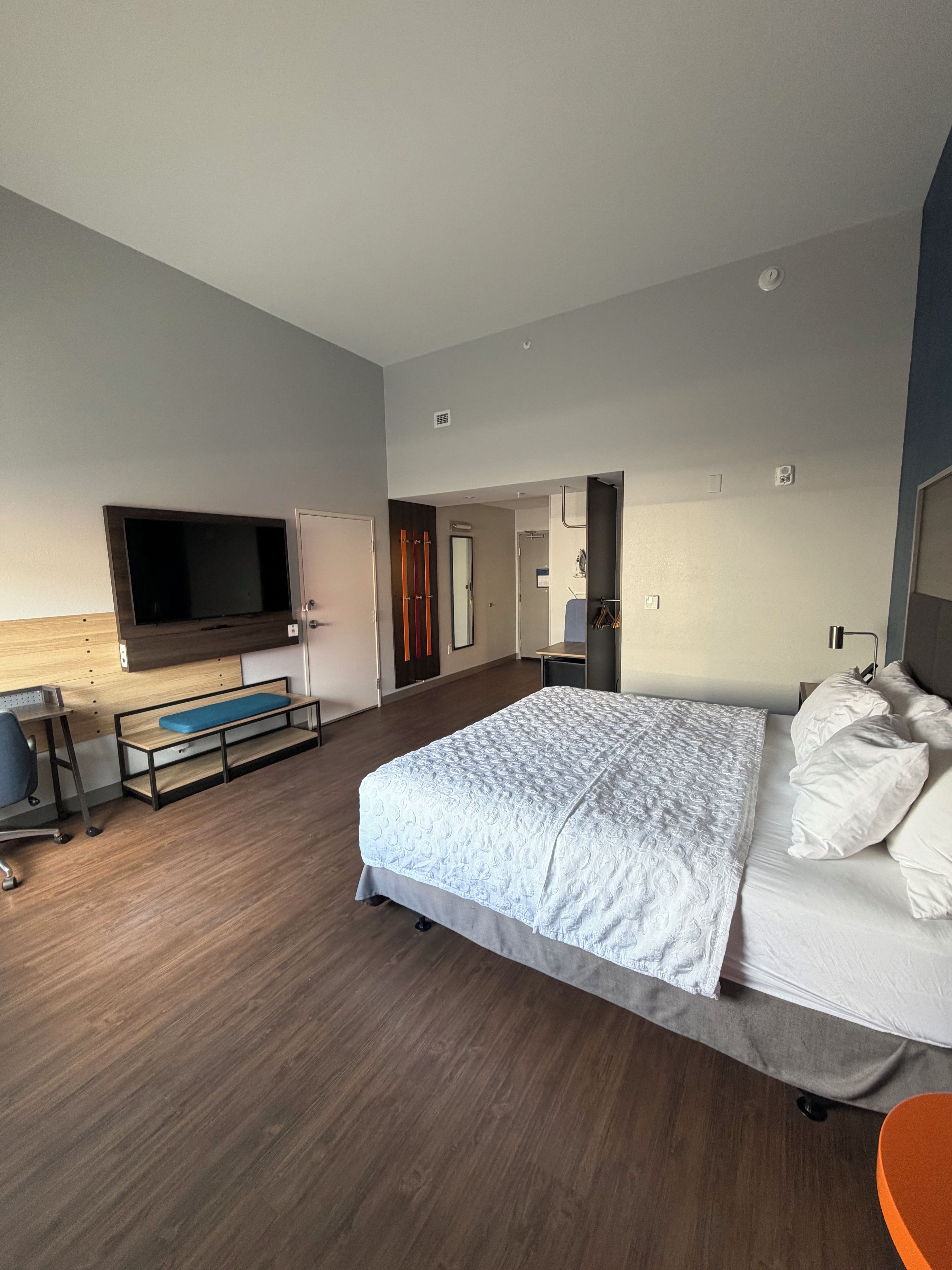 A modern hotel room with a large bed, a flat-screen TV mounted on the wall, a desk with a chair, and a bench.