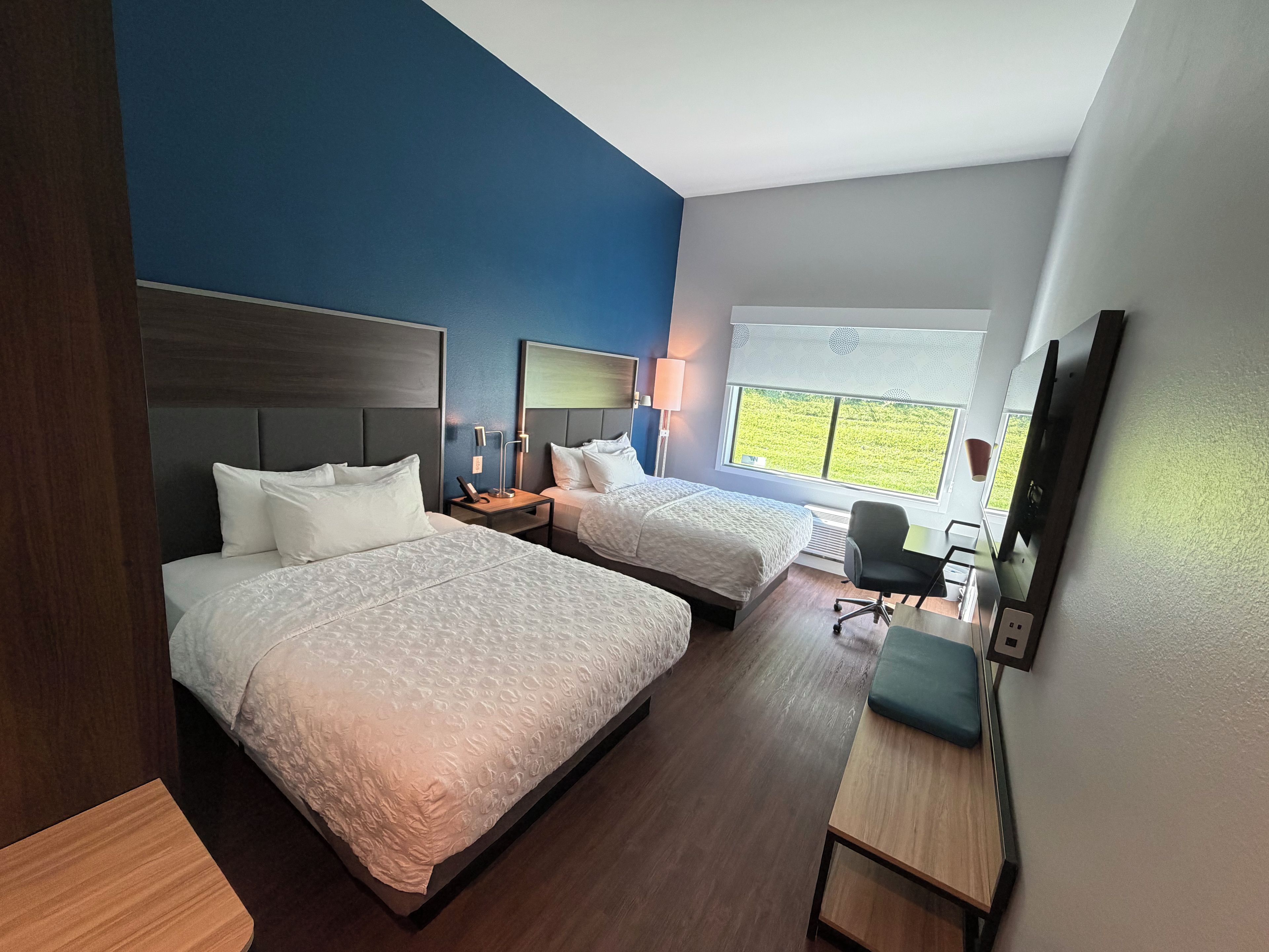 Een hotelkamer met twee bedden, een bureau, een stoel, een televisie en een raam met uitzicht op groen.