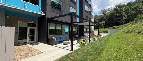 Un hotel moderno con fachada azul, un área de estar al aire libre cubierta y un césped ajardinado.