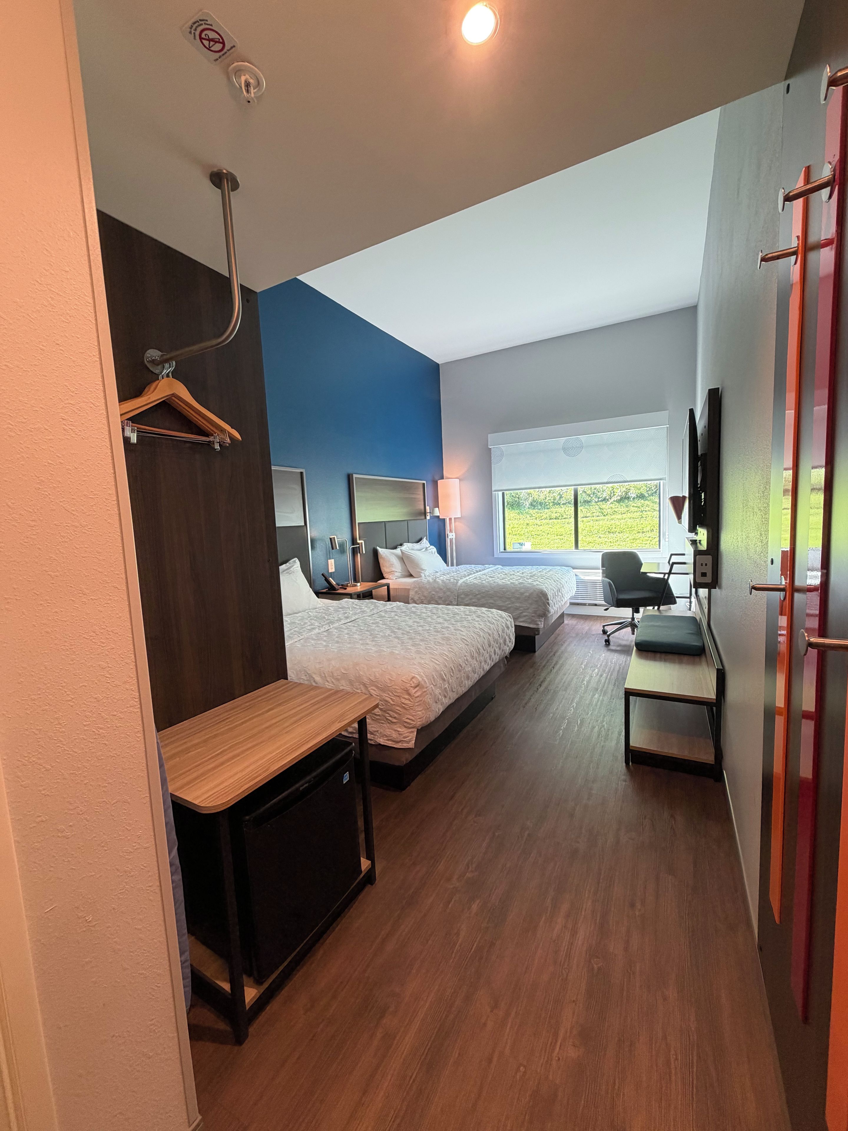 A hotel room with two beds, a desk, a chair, a TV, and a window with a view of greenery.