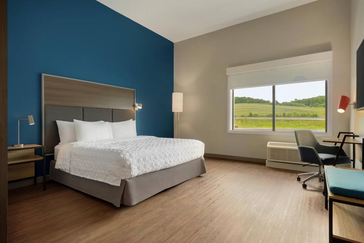 A modern hotel room with a large bed, a desk, a chair, a window with a view of a green landscape, and a wall-mounted lamp.