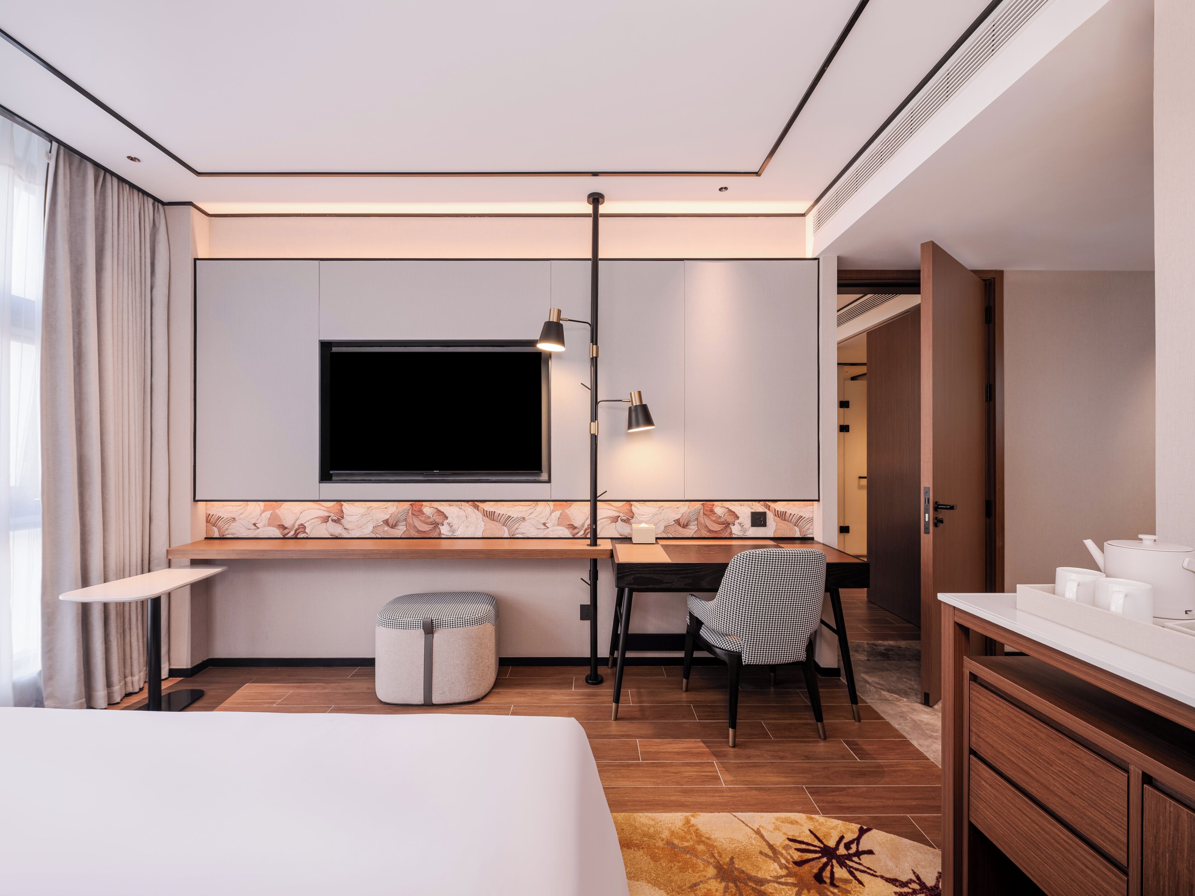A modern hotel room with a bed, a desk with a chair, a television, a lamp, and a bathroom with a sink and mirror.