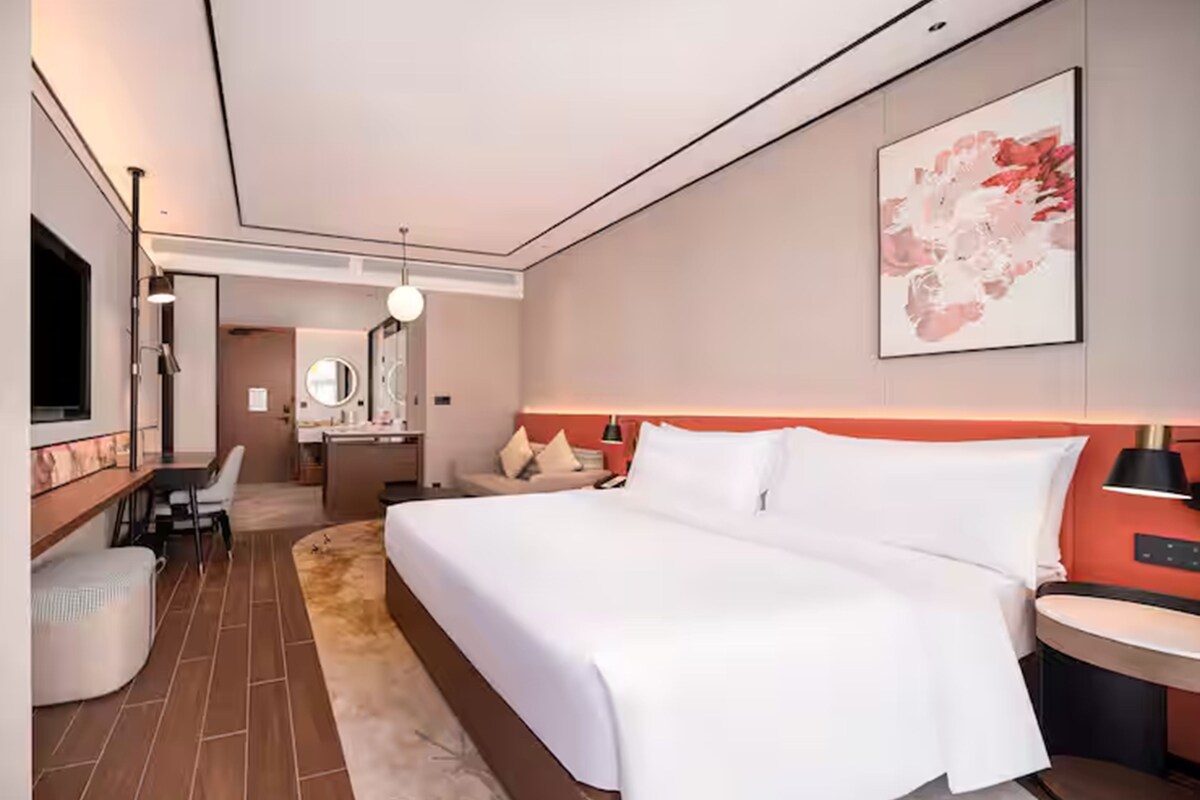 A modern hotel room with a large bed, a desk, a chair, a vanity with a mirror, and a painting on the wall.