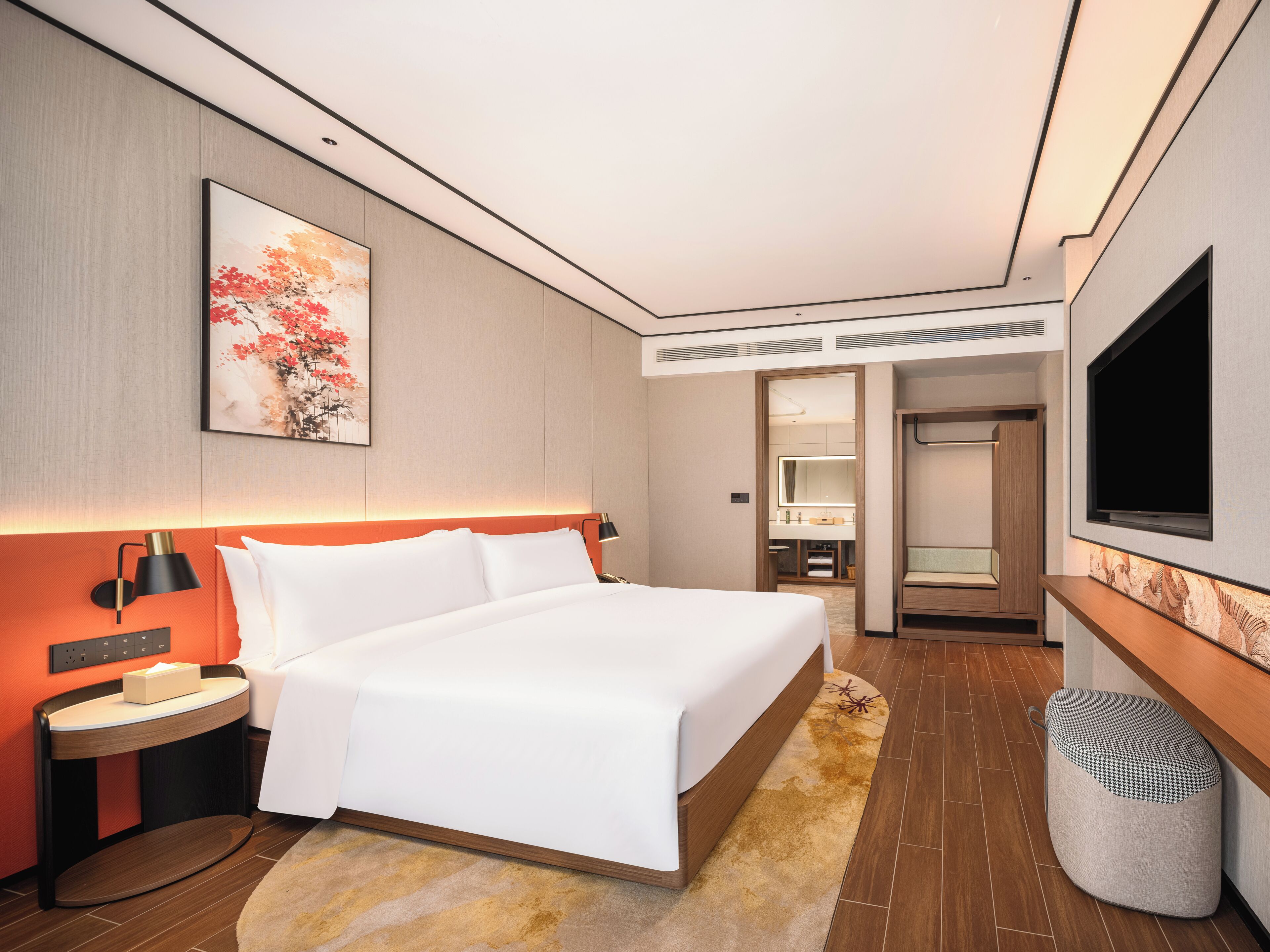 A modern hotel room with a bed, bedside table, lamp, artwork, television, and a bathroom visible through an open door.