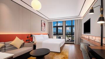 A modern hotel room with a bed, sofa, dining table, and a view of the city through large windows.