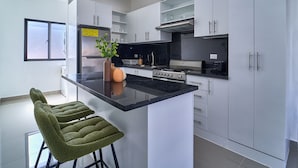 A modern kitchen with white cabinets, a black countertop, and stainless steel appliances. There are two green bar stools and a vase with a plant on the island. - Cozy Nest (La Romana)
