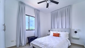 A bedroom with a bed, curtains, a ceiling fan, a window, and a nightstand. - Cozy Nest (La Romana)