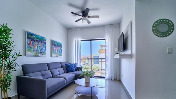 A modern living room with a grey sofa, a small round coffee table, a ceiling fan, and a large window with a view of buildings. - Cozy Nest (La Romana)