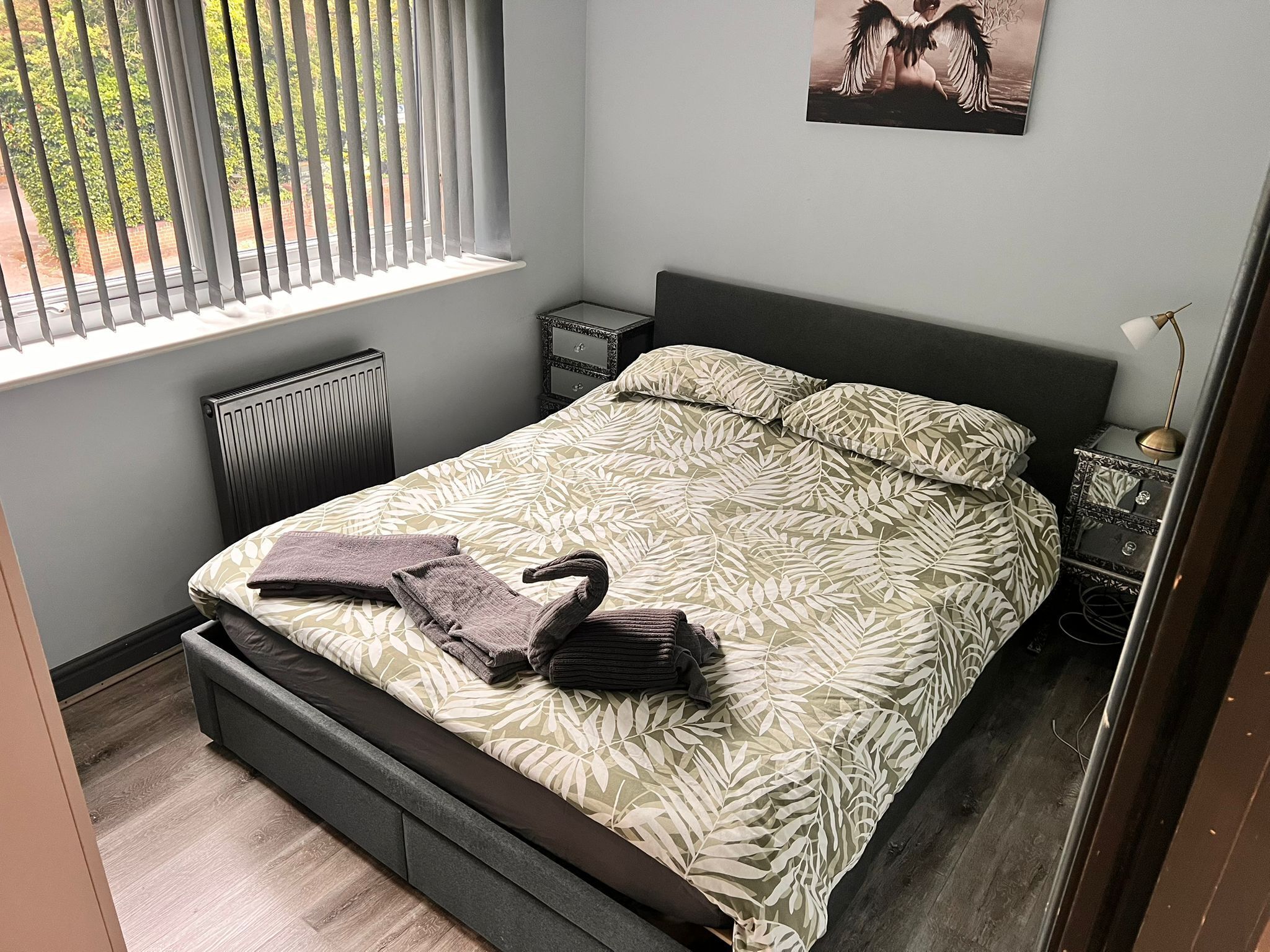 Quarto com cama, mesa de cabeceira, radiador, janela com persianas e quadro pendurado na parede.