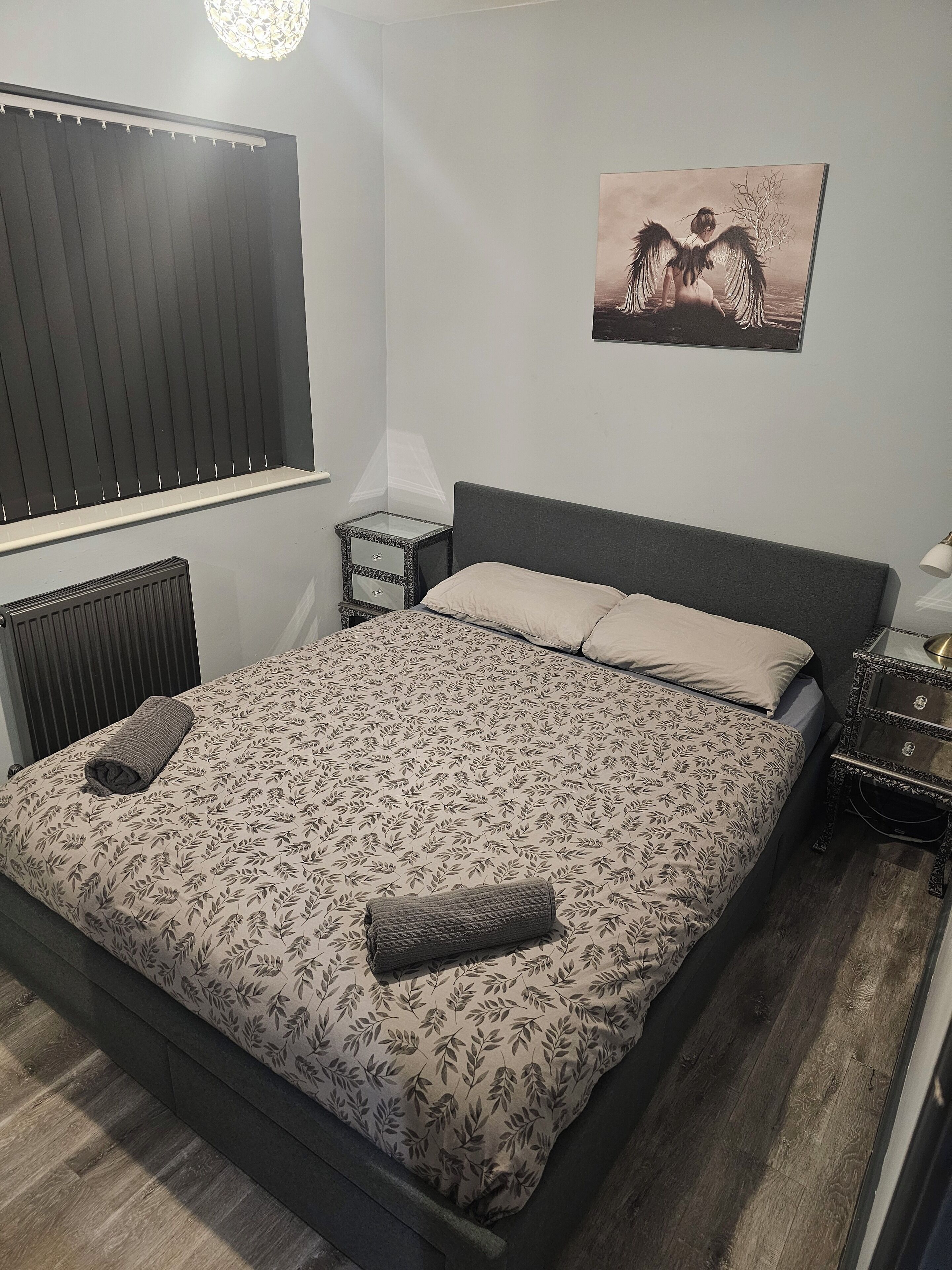 Een netjes opgemaakt bed met een bloemenbedekker, een nachtkastje met een lamp en een aan de muur hangend schilderij.