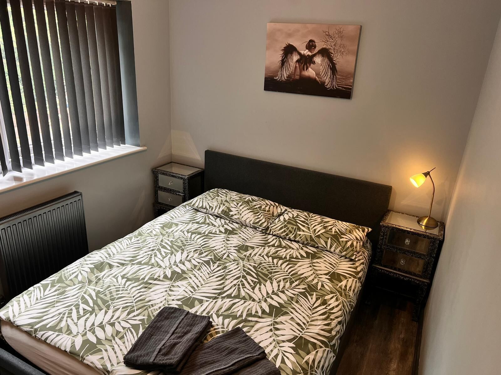 Un dormitorio con cama, mesita de noche, lámpara, radiador y ventana con persianas.